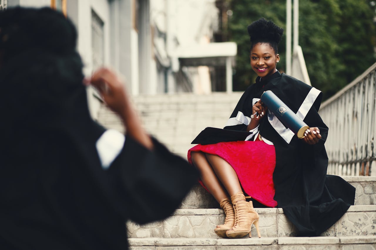 ours-journey Black female graduate in gown smiles with diploma on city stairs, embodying achievement.